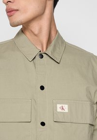 Hemden in hellem Khaki, aus strukturiertem Stoff gefertigt. Verfügt über einen Kragen, zwei Brusttaschen und ein kleines Markenetikett mit rotem Text.
