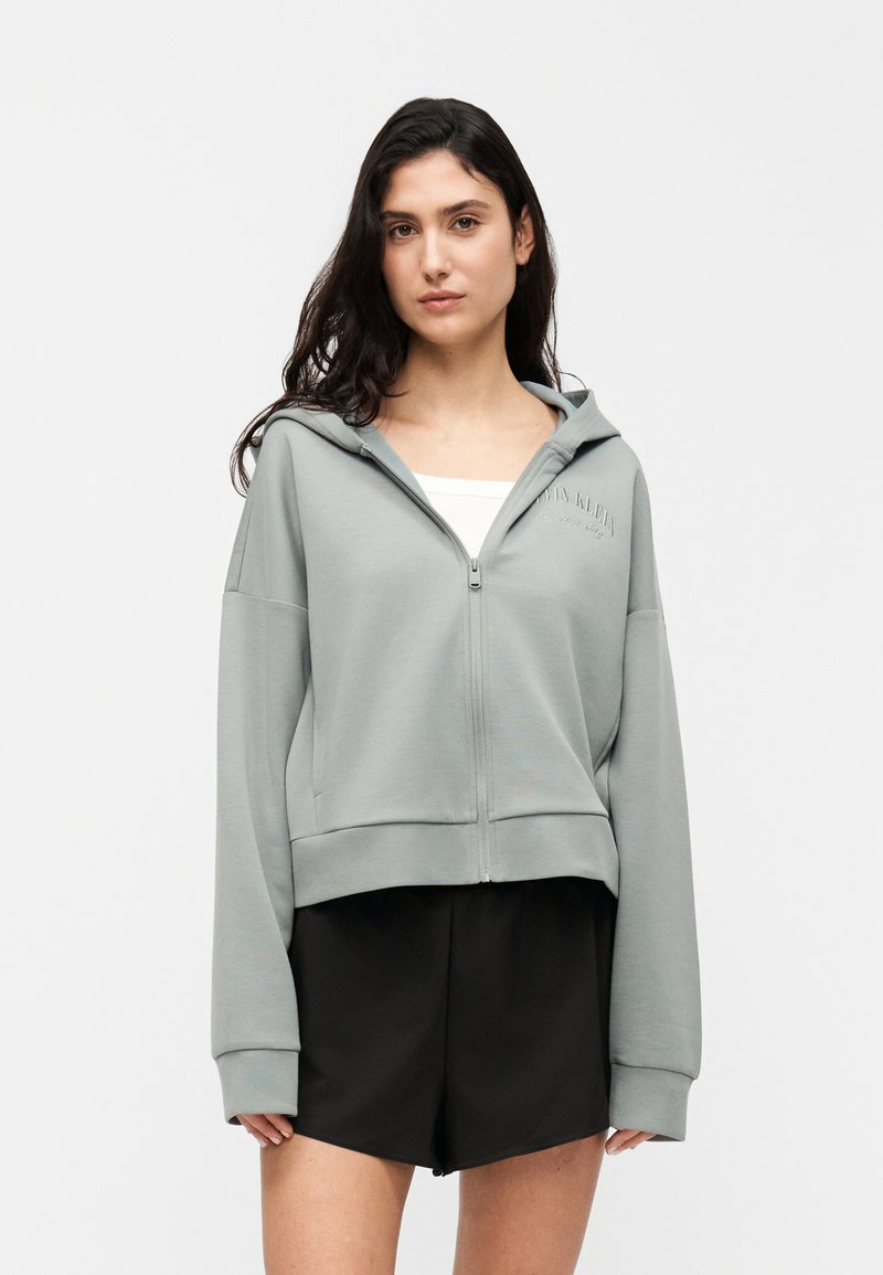 Ανοιχτό πράσινο κοντό φούτερ με φερμουάρ σε απαλό ύφασμα, με κουκούλα, φαρδιά μανίκια και διακριτική λεπτομέρεια λογότυπου· συνδυασμένο με μαύρο σορτς.