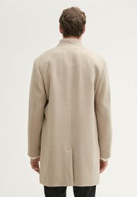 Beige Wollmixmantel mit geradem Schnitt, der minimale Nähedetails und einen hohen Kragen aufweist. Glatte Textur; keine sichtbaren Beschläge.