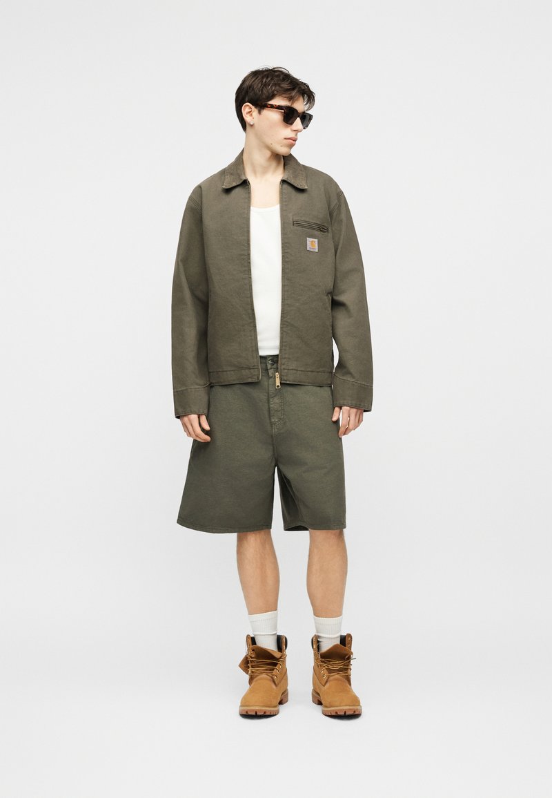 Giovane uomo con occhiali da sole, giacca verde oliva e pantaloncini, canottiera bianca, calzini bianchi e stivali da lavoro color sabbia, in piedi contro uno sfondo neutro.