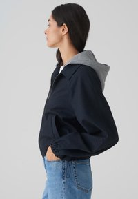 Veste bleu marine avec capuche grise, fermeture éclair et poignets élastiques. Fabriquée en tissu texturé et dispose de poches latérales.
