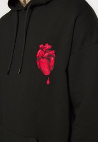 Sudadera negra con corazón anatómico rojo y una sola gota de sangre bordada en el pecho, mostrando parte de un cuello y hombro tatuados.