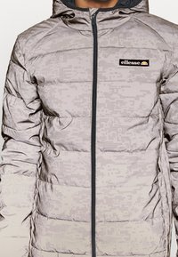 Lysegrå digital camouflage pufferjakke med lynlås foran og sort ellesse-logo på brystet, båret af en person mod en ensfarvet baggrund.