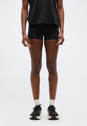 Person iført sorte sportsshorts, sorte sneakers med hvide såler, hvide sokker og en sort top, stående mod en ensfarvet baggrund.