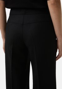 Detailní záběr na osobu oblečenou v černých kalhotách s vysokým pasem, které mají viditelné švy a hladkou texturu látky.