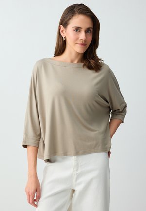 Femme aux cheveux bruns mi-longs portant un haut beige ample à manches 3/4 et un pantalon blanc, debout devant un fond clair uni.