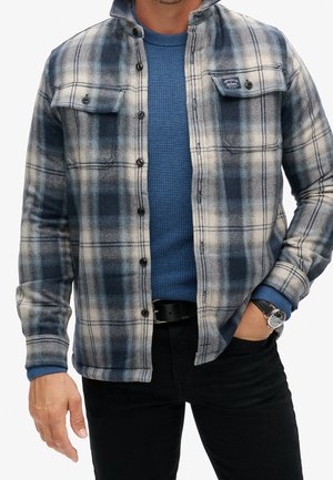 Mann trägt ein blau kariertes Hemd über einem blauen Strickpullover, schwarze Hose, schwarzen Gürtel und Armbanduhr, Hand in der Tasche.