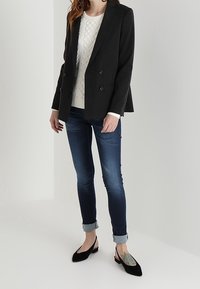 Blazer negro de doble botonadura sobre un suéter blanco de punto trenzado, combinado con vaqueros azul enrollados y zapatos planos negros. Los detalles incluyen un tatuaje en el tobillo.