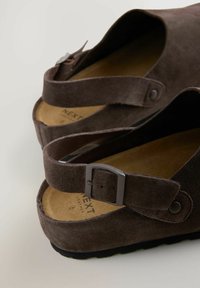 Braune Wildleder-Sandalen mit einem geschwungenen Design, verstellbarem Knöchelriemen mit einer Metall-Schnalle und einer strukturierten Sohle. Weiche Innensohle sichtbar.