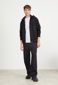 Sudadera con capucha negra y bolsillo frontal, combinada con una camiseta blanca y pantalones negros holgados. Calzado oscuro con detalles texturizados.