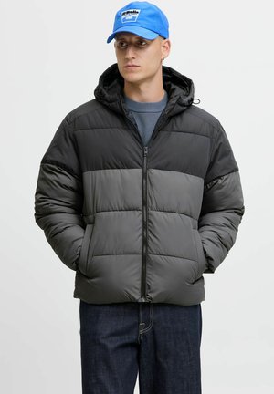 Puffermantel mit einem Farbblockdesign in Schwarz und Grau, Reißverschluss vorne, verstellbare Kapuze und gestepptem Textil; das Modell trägt eine blaue Mütze.