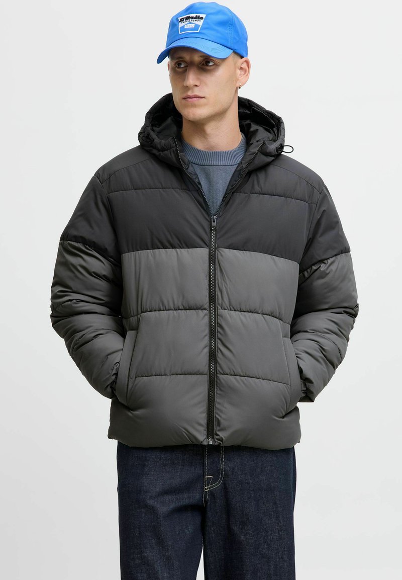 Puffermantel mit einem Farbblockdesign in Schwarz und Grau, Reißverschluss vorne, verstellbare Kapuze und gestepptem Textil; das Modell trägt eine blaue Mütze.