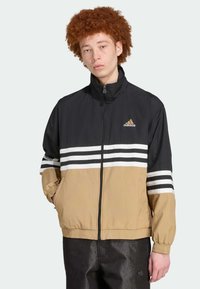 Črn in kašmir windbreaker z visokim ovratnikom, ki ima bele in črne črte preko prsi ter blagovno znamko v zlatem. Lahka tkanina.