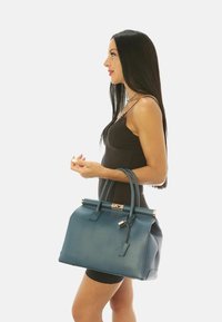 Bolso de mano de cuero verde azulado con forma estructurada, asas dobles y hardware en tono dorado. Presenta un acento de etiqueta y un acabado texturizado.