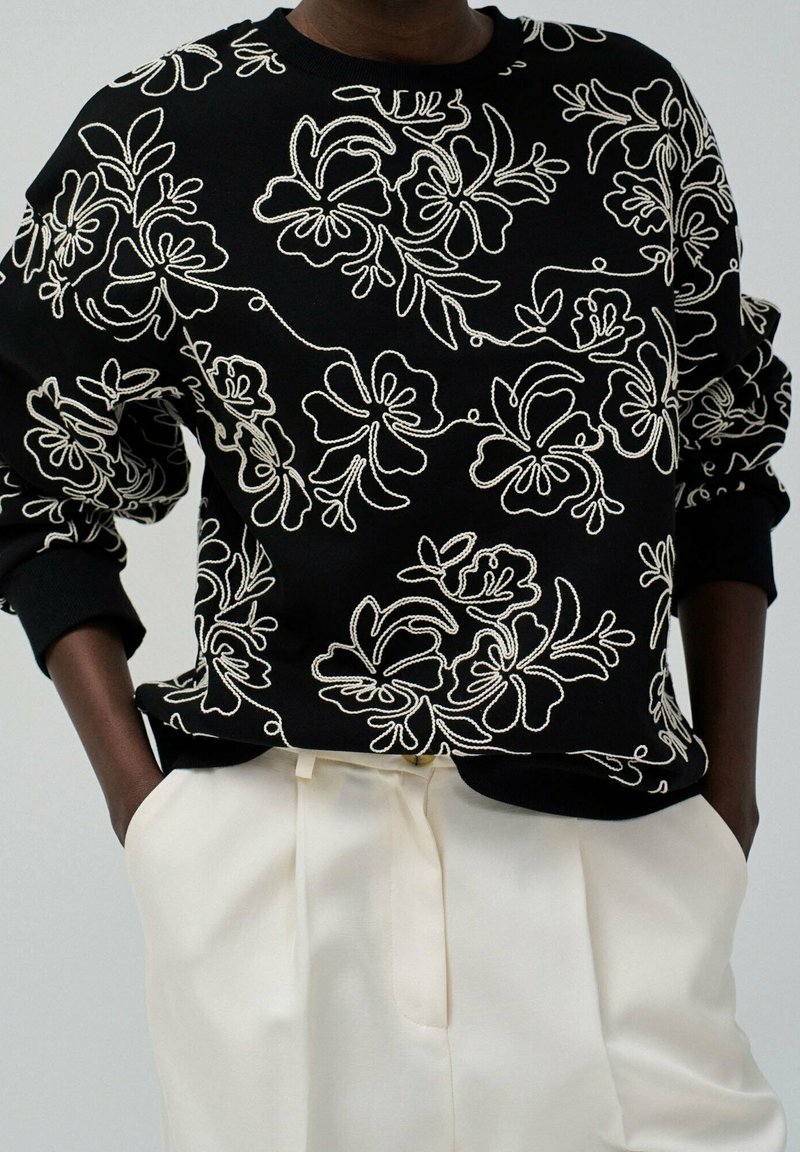 Personne portant un sweat-shirt noir avec une broderie florale blanche et un pantalon crème taille haute, mains dans les poches, sur fond uni.