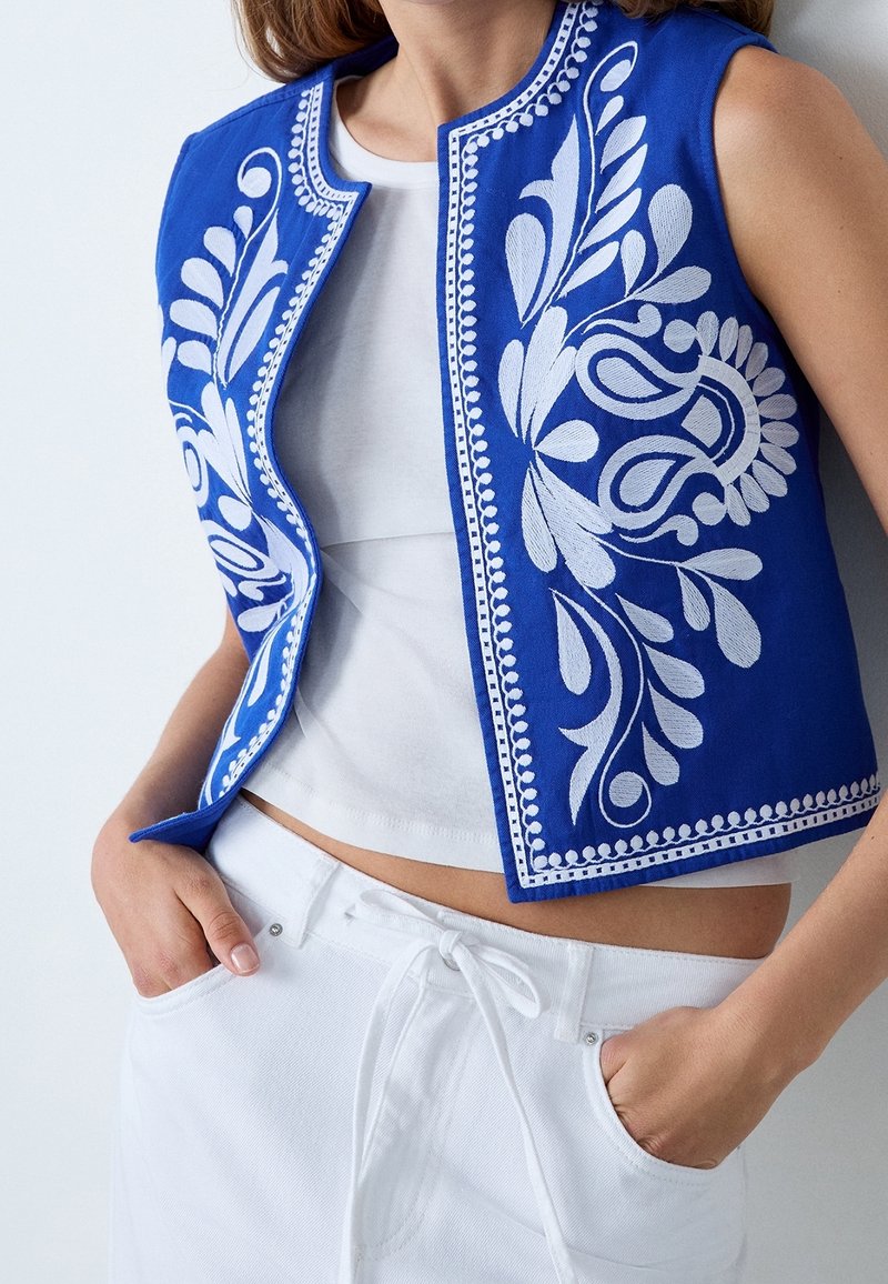 Blå broderet vest med hvide blomsterprint, rund halsudskæring og tætsiddende pasform, båret over en hvid top og kombineret med hvide shorts.