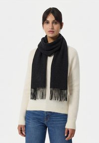 Svart ullsjal med fransiga ändar, som vilar runt halsen. Har en diskret logotyp. Bärs över en cremefärgad tröja och blå jeans.