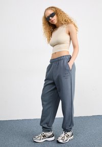 Pantalones de chándal grises con cintura elástica, bolsillos laterales y estampado "NEW BALANCE 1906". Top corto beige, collar de cadena plateada, gafas de sol y zapatillas metálicas.