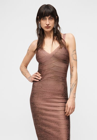 Γυναίκα με μακριά σκουρόχρωμα μαλλιά και τατουάζ, φορώντας εφαρμοστό μεταλλικό φόρεμα σε ροζ χρώμα και κρεμαστά σκουλαρίκια με κρύσταλλα, μπροστά σε απλό φόντο.