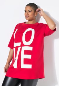 T-shirt rosso oversized con grande testo bianco "LOVE". Maniche corte, scollo rotondo, tessuto morbido. Leggings neri completano il look.