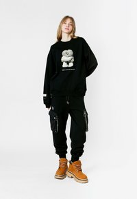 Sudadera negra de gran tamaño con estampado gráfico, combinada con pantalones cargo negros con bolsillos con cremallera, y botas de cordones en color marrón con suelas texturizadas.