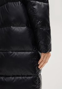 Czarna pikowana kurtka z błyszczącą, przeszywaną fakturą. Posiada poziome przeszycia i boczną kieszeń, prezentując elegancki design i izolację.