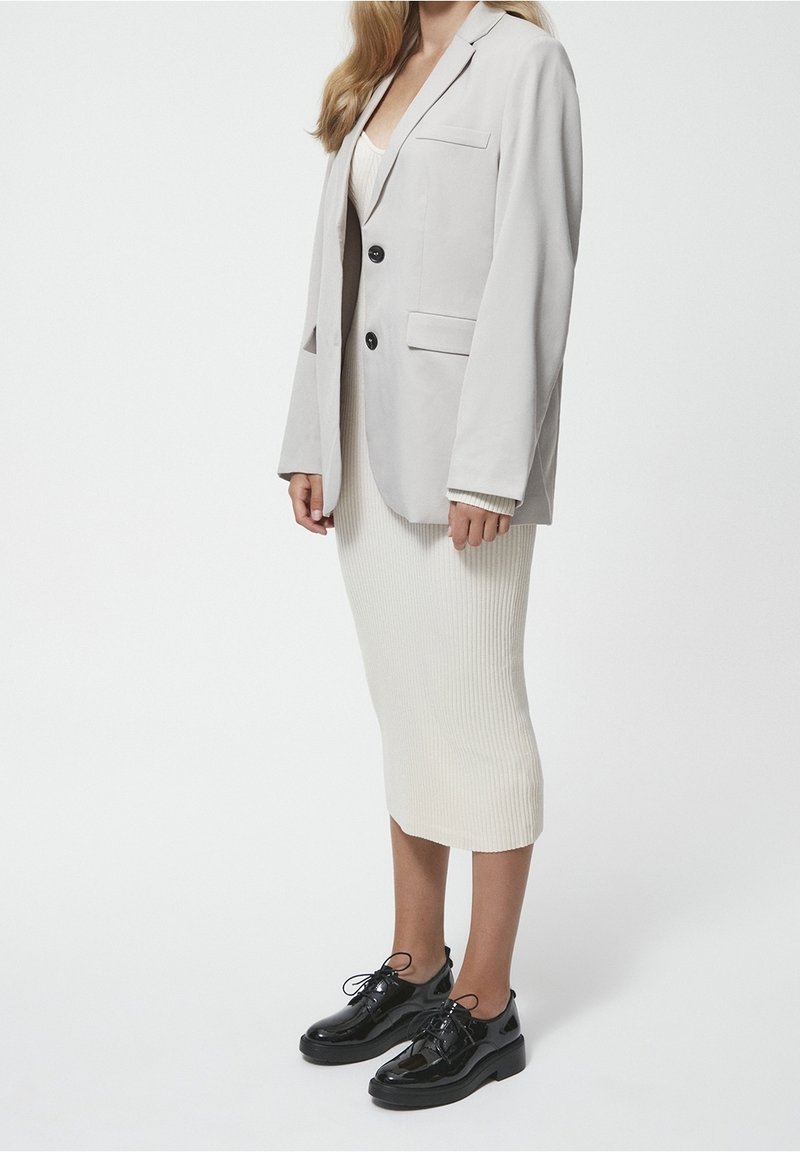Blazer gris avec deux boutons noirs et poches avant porté sur une robe crème côtelée. Chaussures noires en cuir verni à lacets complètent le look.