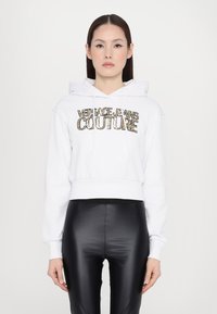 Hoodie bianco corto con cappuccio e lacci, decorato con lettering dorato che recita "Versace Jeans Couture". Abbinato a leggings neri lucidi.