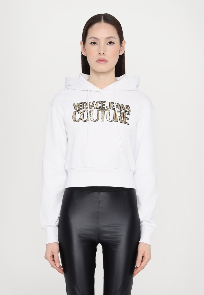 Hoodie bianco corto con cappuccio e lacci, decorato con lettering dorato che recita "Versace Jeans Couture". Abbinato a leggings neri lucidi.