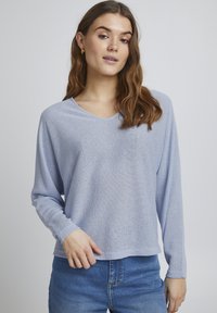 Bluză lungă de culoare albastru deschis, dintr-un material texturat, cu decolteu în V și croială lejeră, purtată împreună cu blugi cu talie înaltă.