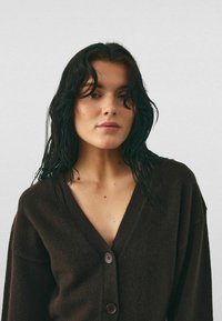 Frau mit nassen schwarzen Haaren, die einen dunkelbraunen, geknöpften Cardigan vor einem einfarbigen hellen Hintergrund trägt.