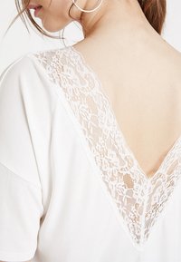 Bílé tričko s hlubokým výstřihem do V, zdobené jemným bílým krajkovým lemem na zádech, z hladké látky a s krátkými rukávy.