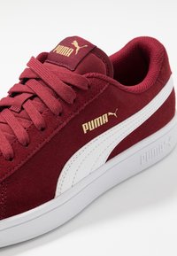 Bordové semišové tenisky Puma s bílým bočním pruhováním, zlatými logy Puma na jazyku a boku, a bílou gumovou podrážkou.