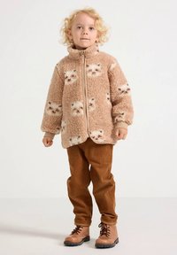 Brun fleecejacka med kattmönster, dragkedja fram och hög krage. Kombinerad med bruna manchesterbyxor och bruna stövlar.