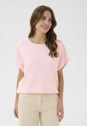 Naine, kellel on pikk laineline pruunid juuksed, kandes pärlikõrvarõngaid, heleroosa lühikeste varrukatega ülaosa ja beeži tekstuuriga pükse, naeratab ja vaatab paremale.