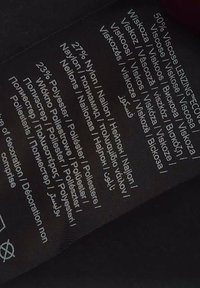 Černý štítek s bílým textem uvádějícím složení tkaniny: 50% viskóza, 27% nylon, 23% polyester; zahrnuje vícejazyčné pokyny k údržbě.