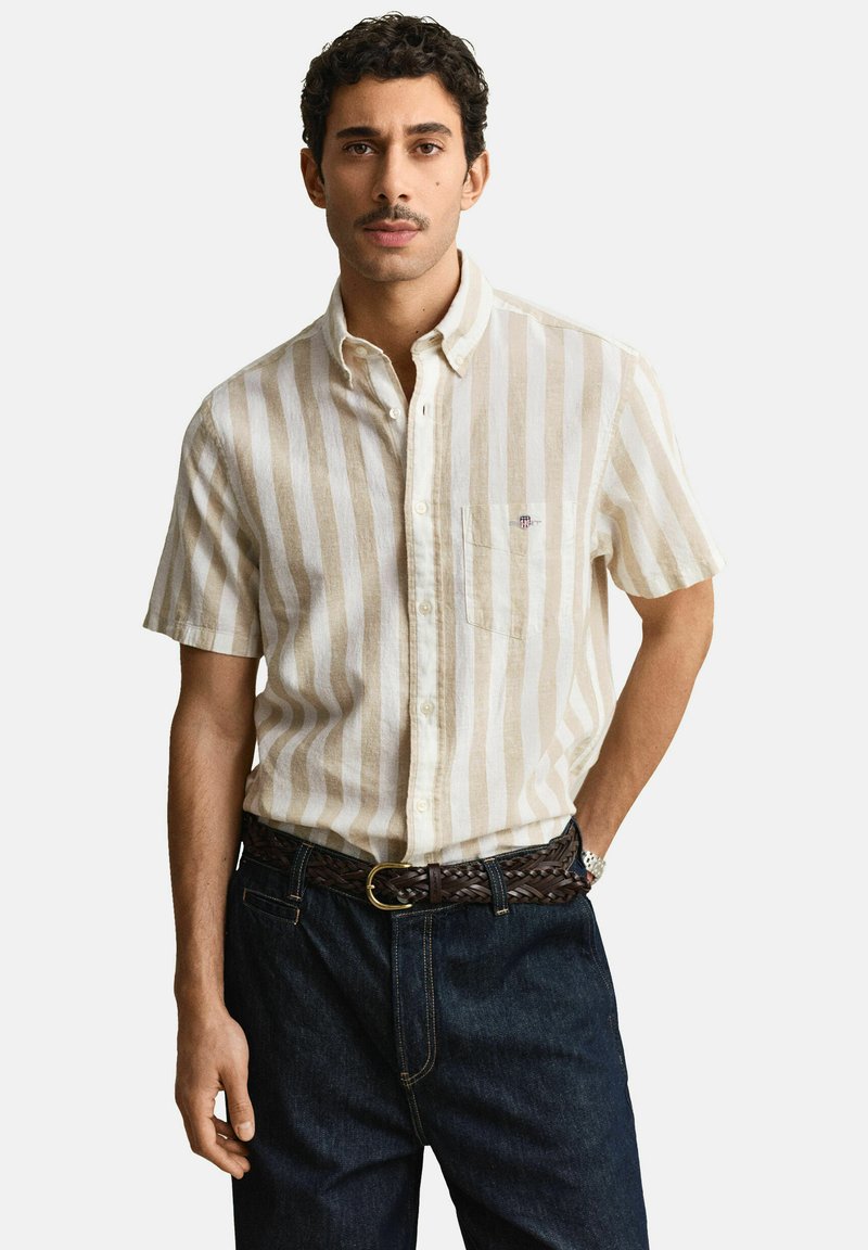 Mann mit dunklem lockigem Haar und Schnurrbart, der ein beige-weiß gestreiftes Kurzarmhemd, dunkle Jeans und einen geflochtenen braunen Gürtel trägt, steht.