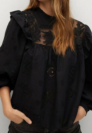 Femme portant un chemisier noir à manches bouffantes, broderie en dentelle florale et panneau supérieur transparent avec détails en perles, mains dans les poches.