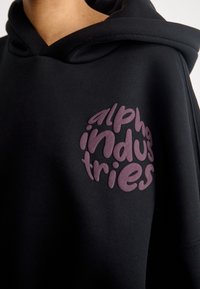 Sweat à capuche noir avec logo "Alpha Industries" texturé violet sur la poitrine, montrant le haut du torse et une partie de la capuche.