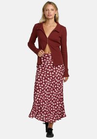 Veste zippée côtelée marron accompagnée d'une jupe bordeaux à motifs floraux, longue jusqu'aux chevilles. Des chaussures de sport noires complètent le look.