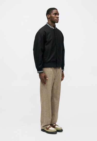 Μαύρο μπουφάν bomber με ριγέ γιακά, μπεζ παντελόνι από μάλλινο ύφασμα και παπούτσια από μεικτά υλικά με πράσινη λεπτομέρεια και καφέ στοιχεία.