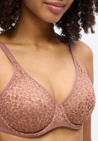 Γυναίκα που φοράει μωβ δαντελένιο σουτιέν με μπανέλα και ρυθμιζόμενες τιράντες, εμφανίζοντας τους ώμους, τον λαιμό και ένα μέρος του προσώπου με σγουρά μαλλιά.