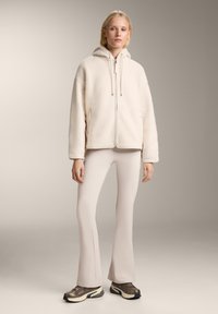 Giacca in pile color crema con cappuccio, chiusura zip e cordini, abbinata a pantaloni svasati beige chiaro e sneakers scure e robuste.