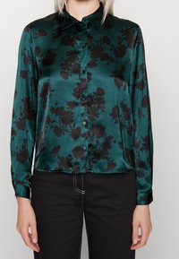 Blusa in satin verde con motivo floreale nero, maniche lunghe, chiusura a bottoni e colletto. Texture liscia con una finitura leggermente lucida.