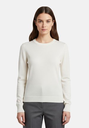 Donna in piedi contro uno sfondo bianco che indossa un maglione bianco a maniche lunghe con scollo rotondo e pantaloni grigi, rivolta in avanti con le braccia lungo i fianchi.