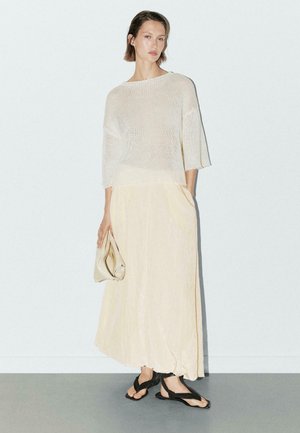 Top leggero in maglia di crema con maniche ampie, abbinato a una maxi gonna fluida di un giallo pallido. Completato con sandali neri flip-flop e una borsa beige.