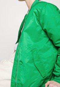 Giacca bomber verde in tessuto lucido con chiusura a zip, colletto a coste e tasche laterali, caratterizzata da una texture liscia.
