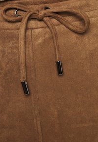 Tissu en daim marron avec une texture lisse, présentant une fermeture par cordon de serrage ornée d'aglets métalliques. Vue en gros plan des détails du matériau.