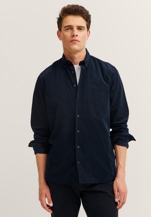 Mann, der ein dunkelblaues Hemd mit Knöpfen und hochgekrempelten Ärmeln über einem weißen Unterhemd trägt, vor einem einfarbigen Hintergrund steht.