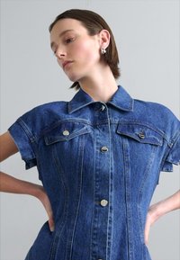 Rochie din denim cu mâneci scurte, culoare albastru deschis, guler, două buzunare pe piept și închidere cu nasturi pe față. Textura pare netedă și bine croită.